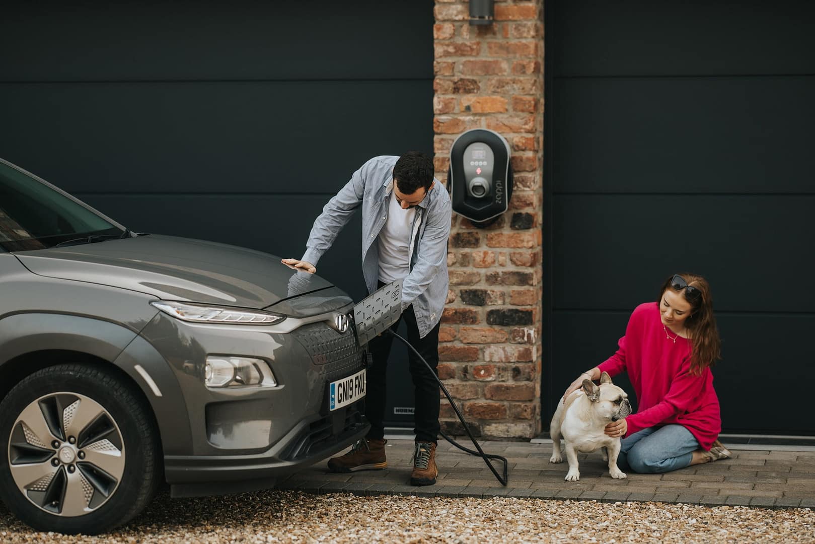 man plugging in zappi ev charger by myenergi  into car, with woman in pink jumper patting a dog
